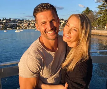 A smiling couple embraces on a sunny waterfront with boats and buildings in the background.