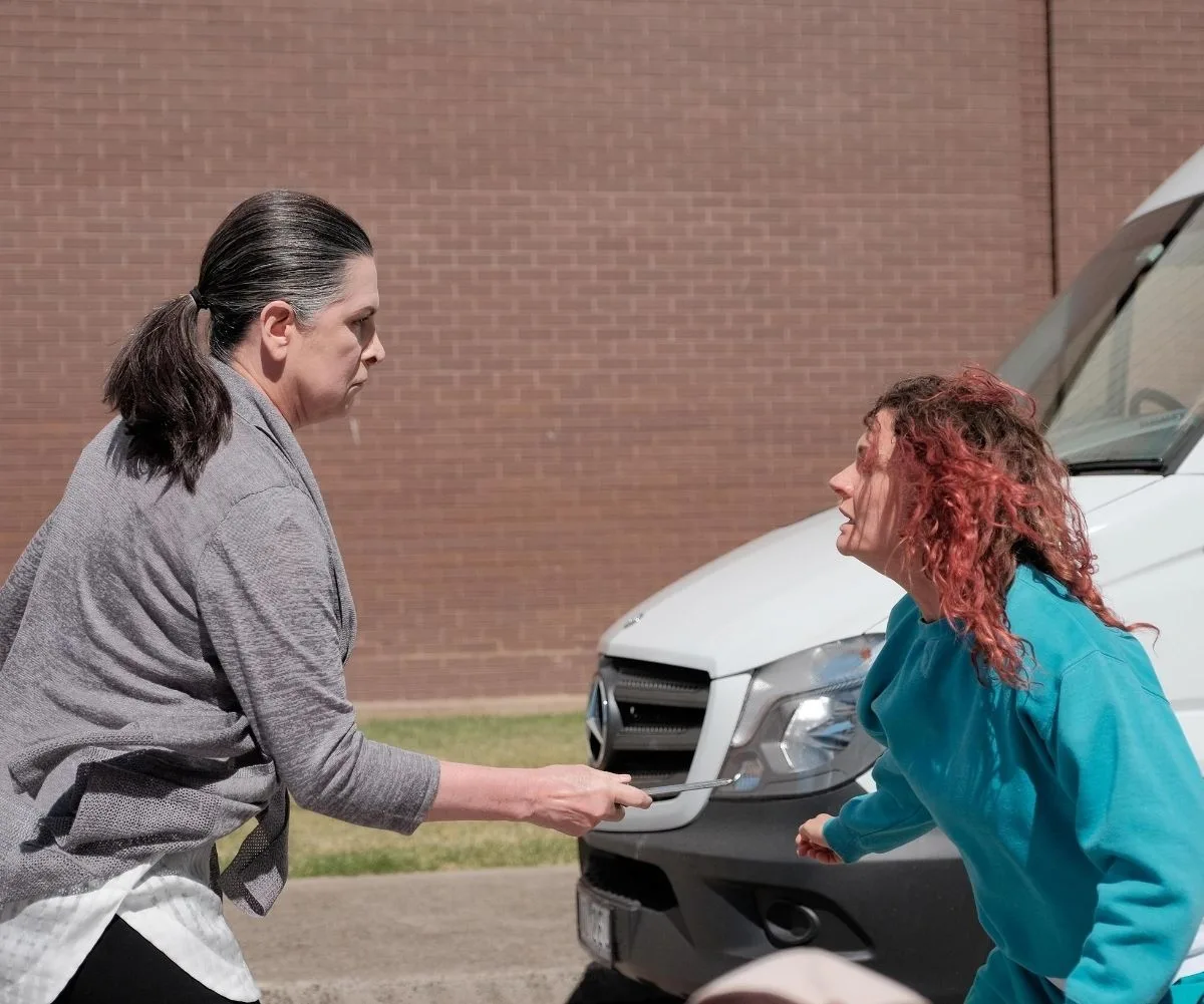 Two women face each other intensely; one holds a sharp object near a van on a sunny day.