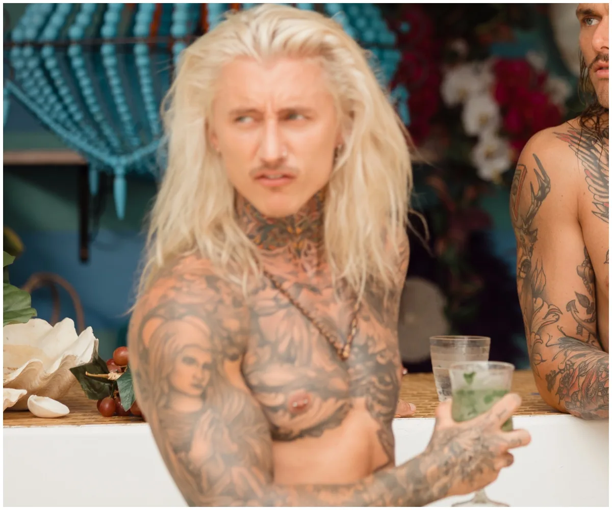 Tattooed person with long blonde hair holds a drink, standing at a bar with colorful decor in the background.