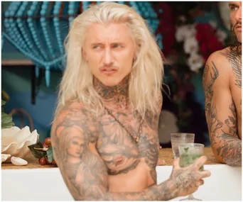 Tattooed person with long blonde hair holds a drink, standing at a bar with colorful decor in the background.