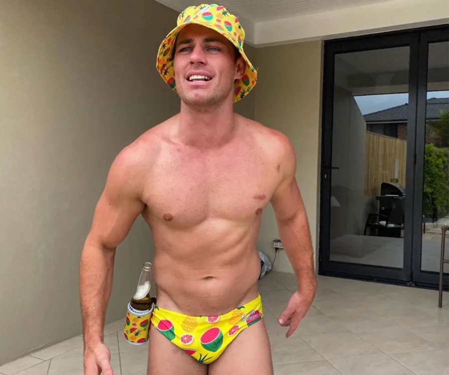 Man wearing colorful swim trunks and bucket hat, holding a drink, standing on a patio.