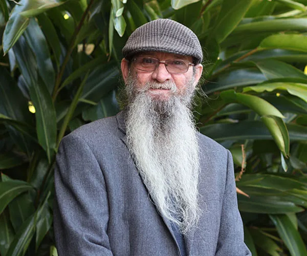 Man with long beard and hat, wearing glasses and a suit jacket, standing in front of large green leaves.