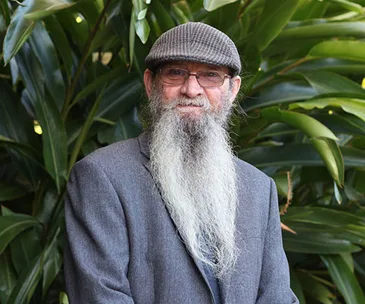 Man with long beard and hat, wearing glasses and a suit jacket, standing in front of large green leaves.