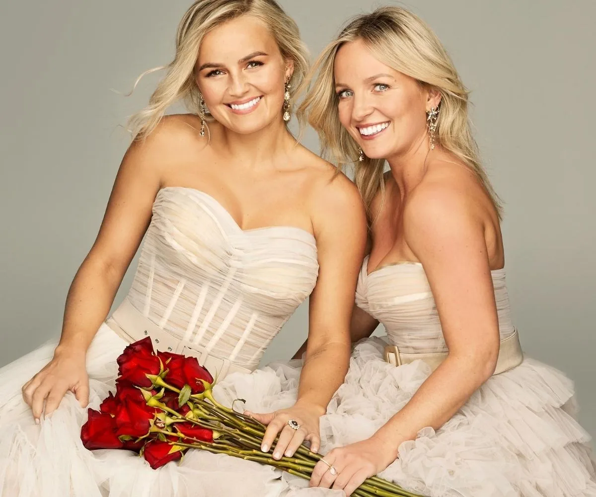 Two women in white dresses, holding red roses, and smiling at the camera.