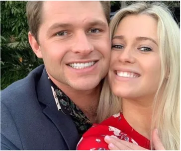 A couple smiling, with the woman showing an engagement ring, embraced and happy outdoors.