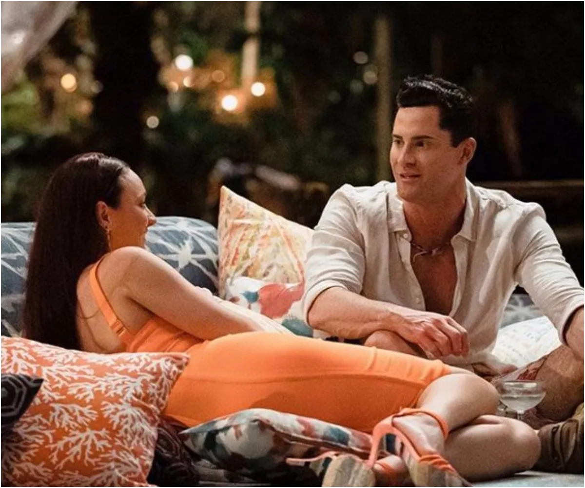 A man and woman lounging on patterned pillows, engaged in conversation in a warmly lit, outdoor setting.