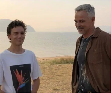 Two men stand on a beach with a calm sea in the background, smiling at the camera.