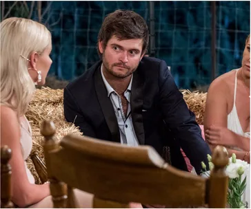 Man in suit sitting on hay with two women during a farmer-themed event, engaged in conversation.