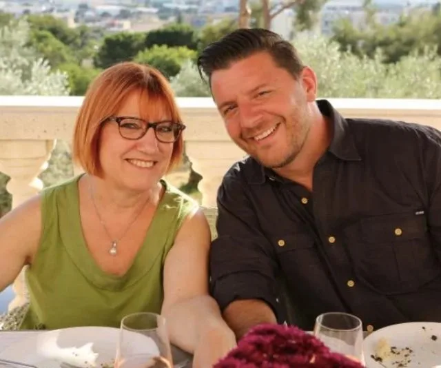 A smiling woman in glasses and greets a smiling man outdoors; meal on table in a sunny setting.