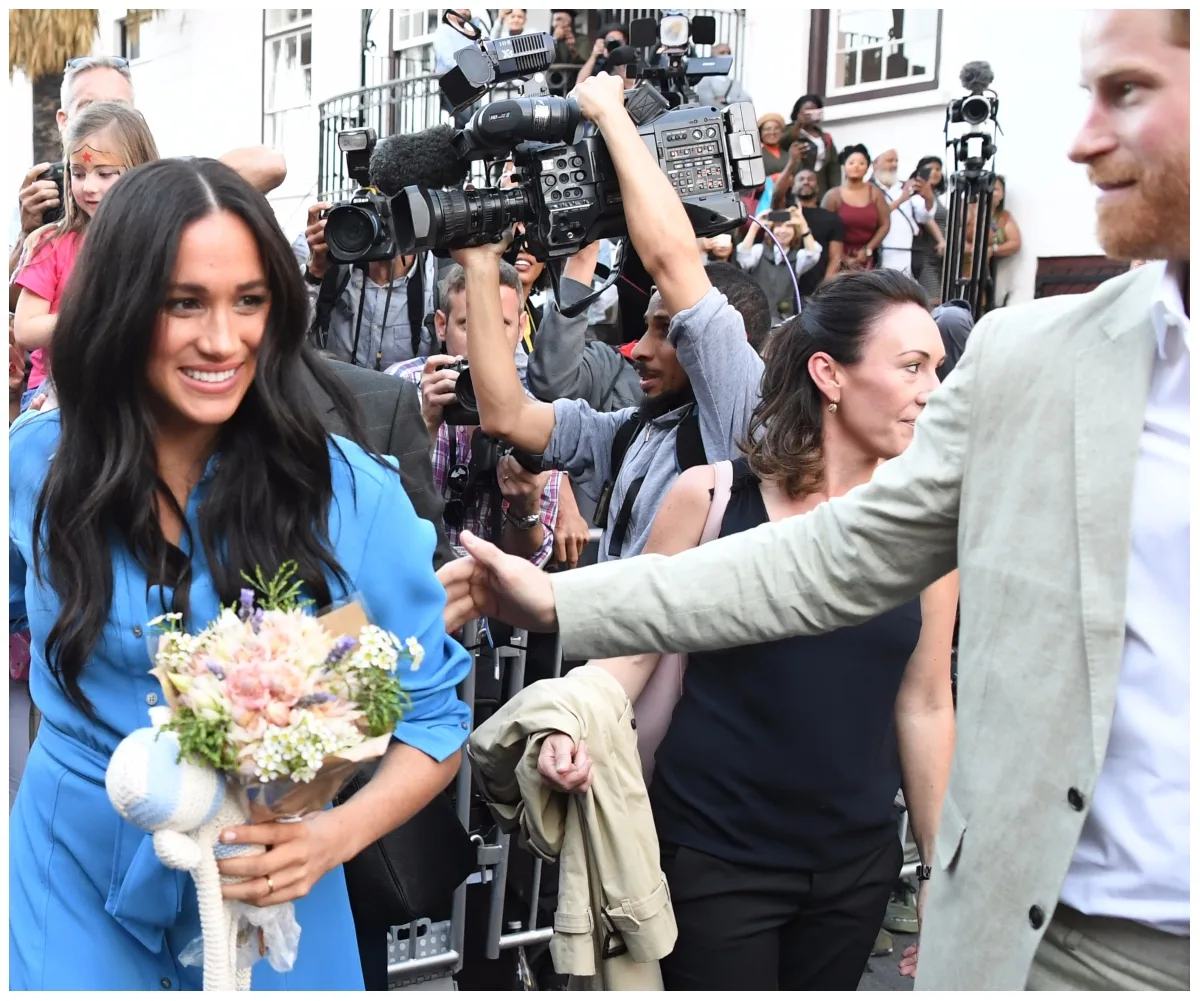 Woman in blue dress holding flowers, smiling amidst a crowd with cameras and people surrounding her.