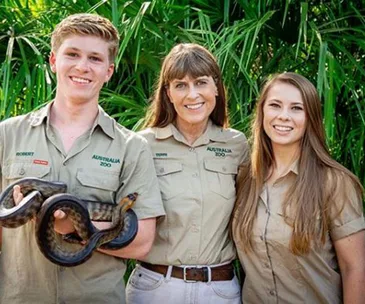 Three people in Australia Zoo uniforms, the left person holding a snake, standing in front of greenery.