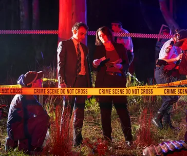 Detectives and police officers at a night crime scene with "Do Not Cross" tape in a wooded area.
