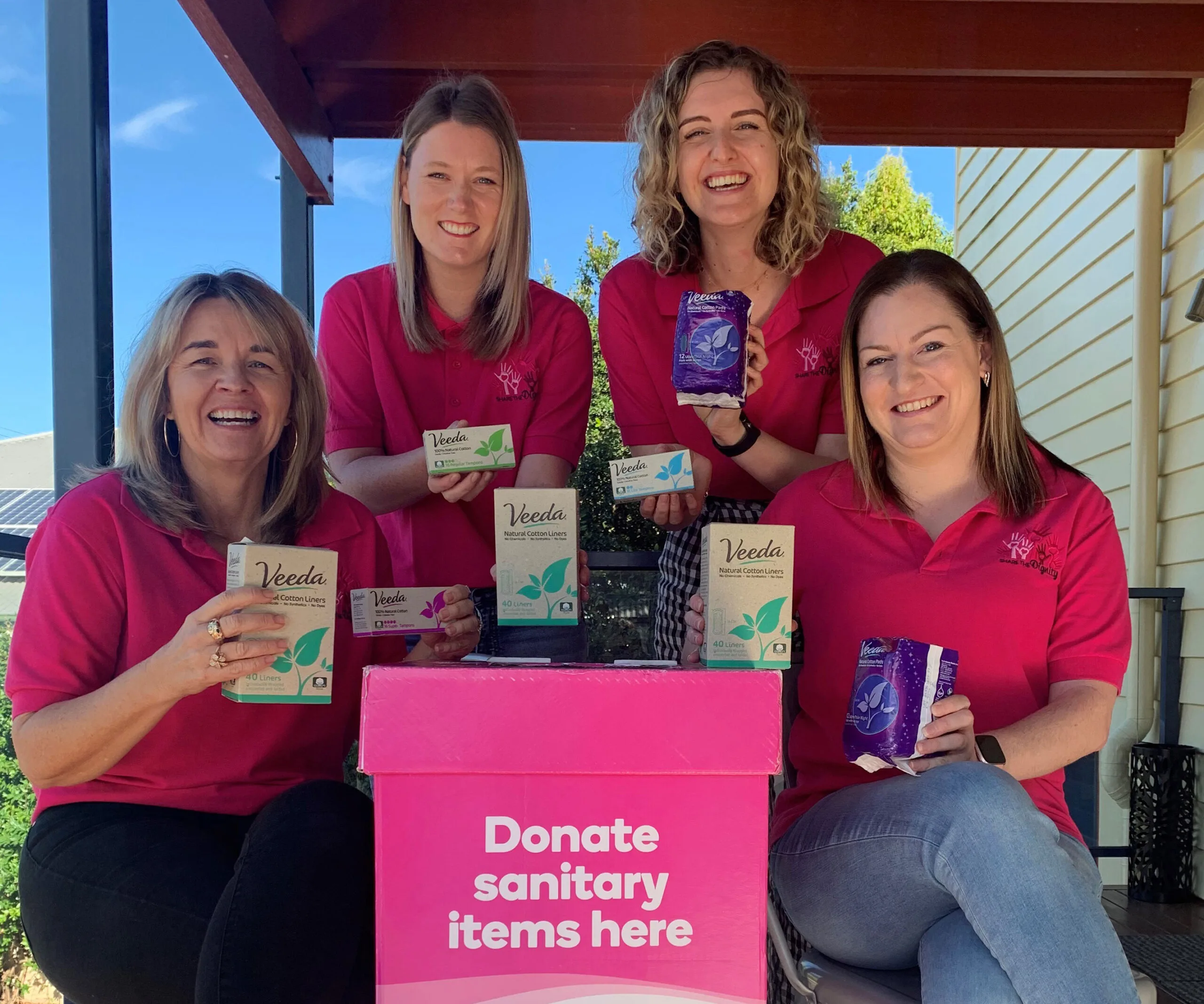 Women in pink shirts holding Veeda sanitary products next to a donation box labeled "Donate sanitary items here."