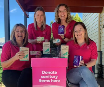 Women in pink shirts holding Veeda sanitary products next to a donation box labeled "Donate sanitary items here."