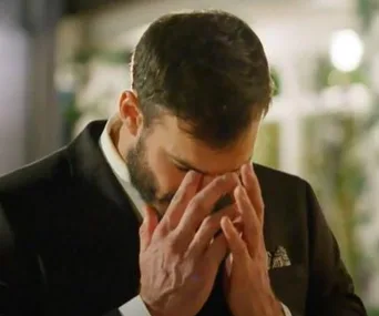 Man in a suit looking stressed, holding his face during a dramatic moment on a reality show.
