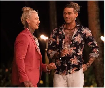 Two men with tattoos, one in a pink suit and the other in a floral shirt, conversing at an outdoor evening event.