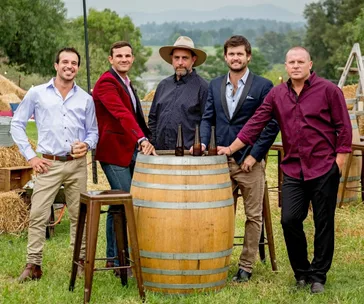 Five men standing around a barrel with bottles in an outdoor rustic setting, wearing varied casual outfits.