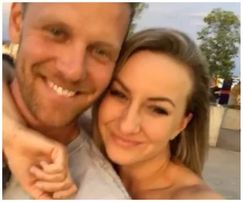 A smiling couple takes a close-up selfie outdoors with trees in the background.