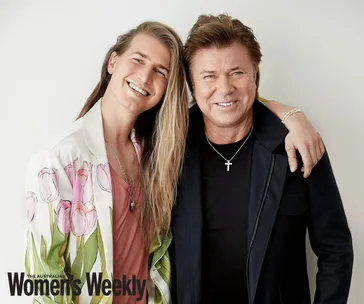 "Two smiling individuals, one with long hair in a floral jacket, the other in black with a cross necklace."