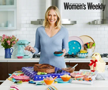 Woman in a kitchen with a chocolate cake, colorful dishes, and flowers, preparing cookies. Australian Women's Weekly cover.