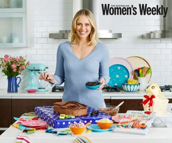 Woman in a kitchen with a chocolate cake, colorful dishes, and flowers, preparing cookies. Australian Women's Weekly cover.