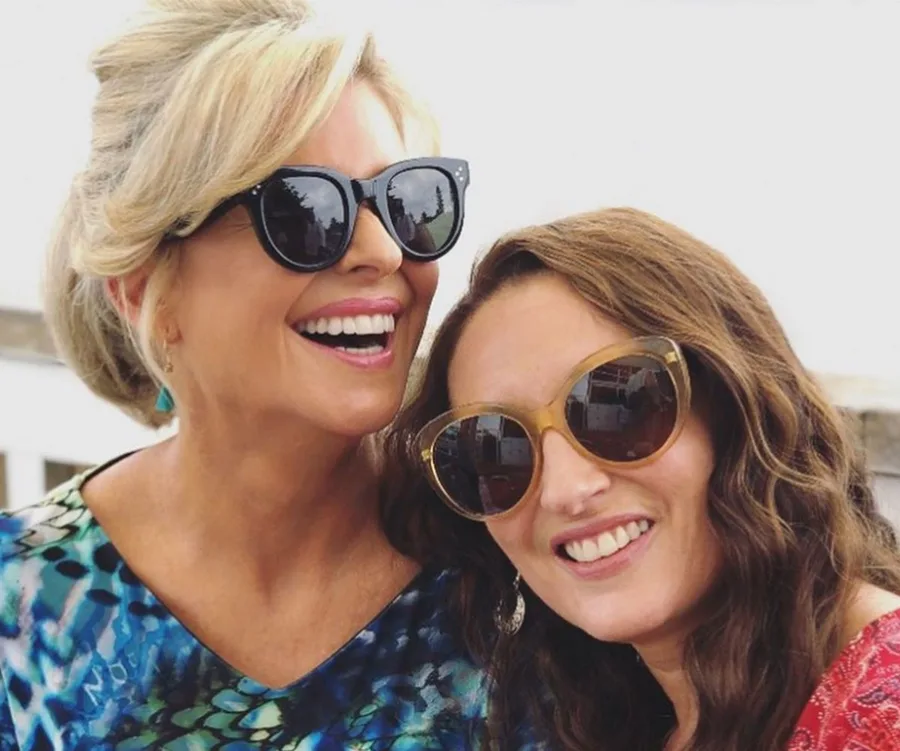 Two women smiling, wearing sunglasses and colorful outfits, outdoors in natural light.