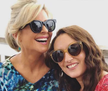 Two women smiling, wearing sunglasses and colorful outfits, outdoors in natural light.