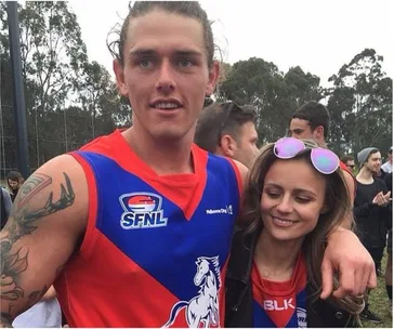Young man in sports jersey with arm around a smiling woman, outdoors with people and trees in the background.