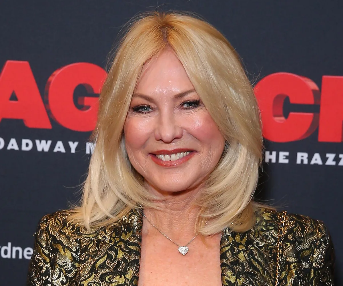 Blonde woman smiling at a red carpet event in a patterned outfit against a poster backdrop.