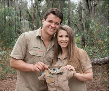 Couple smiling and holding a small khaki shirt with "Australia Zoo" patches in a forest setting.