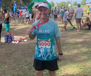 Man holding a medal and wearing a race bib, standing outdoors at a running event with others in the background.