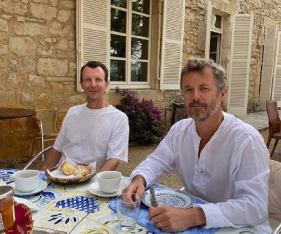 Two men in white shirts sitting at an outdoor table with coffee cups and pastries, in front of a stone building.