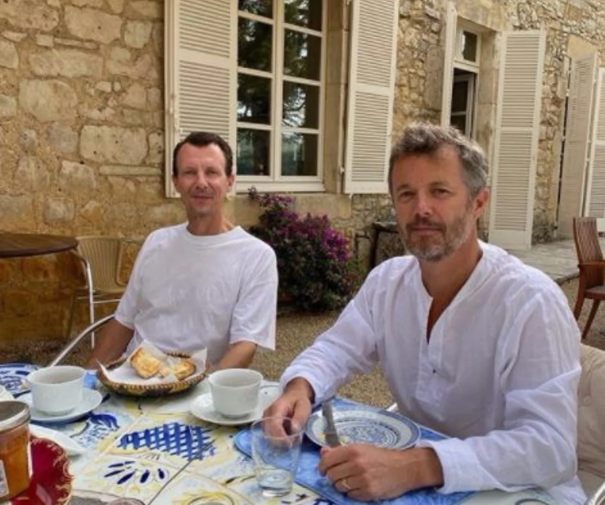 Two men in white shirts sitting at an outdoor table with coffee cups and pastries, in front of a stone building.