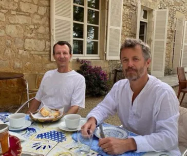 Two men in white shirts sitting at an outdoor table with coffee cups and pastries, in front of a stone building.