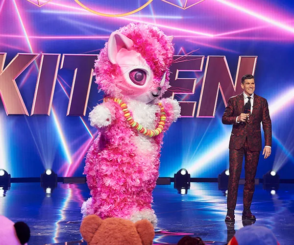 Pink Kitten costume on stage with host in patterned suit, "Masked Singer Australia" in background.