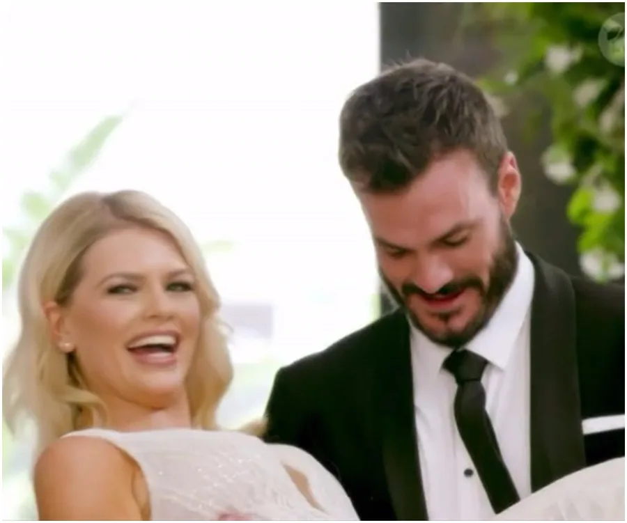 A woman in a white dress is being carried by a man in a suit. They are both smiling, surrounded by greenery.