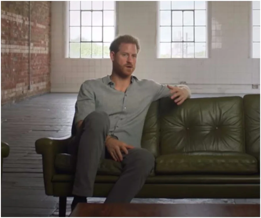 Prince Harry sitting on a green sofa in a spacious room with large windows in a scene from his Netflix documentary.