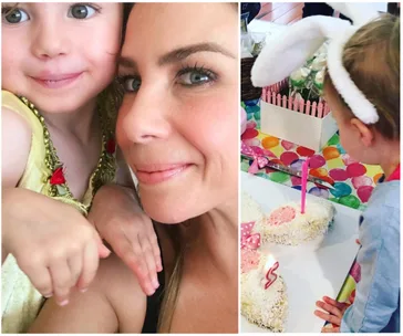 Close-up of a woman and a child smiling; child in bunny ears near a cake with a pink candle on a colorful table.