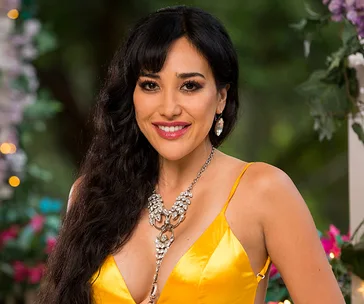 A woman in a yellow dress with long black hair and a jeweled necklace smiles outdoors with floral background.