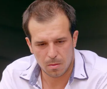 Man looking down with a serious expression, wearing a white shirt with dark collar detail.