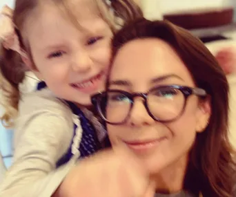 A woman with glasses smiling and hugging a young child with pigtails and a hair bow.