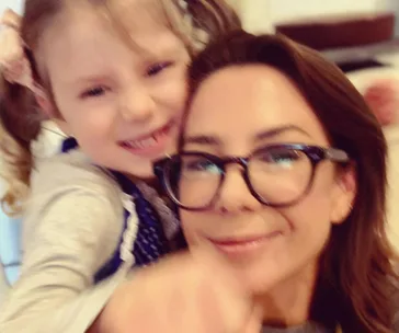 A woman with glasses smiling and hugging a young child with pigtails and a hair bow.
