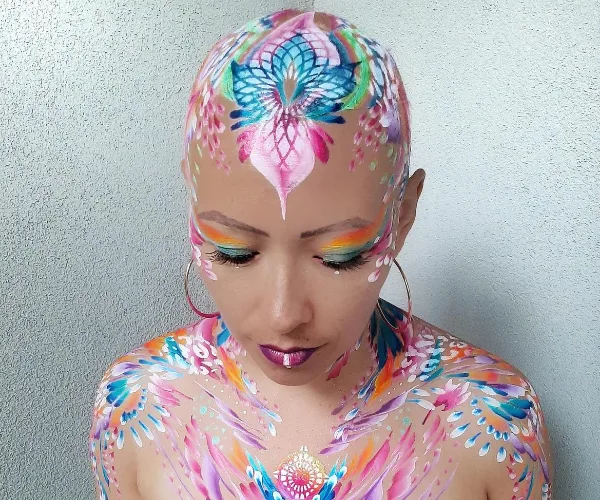 Person with intricate floral body paint design on head and chest, vibrant makeup, looking down.