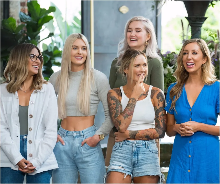 Women from "The Bachelor Australia 2020" stand smiling together, casual outfits, green plants in the background.