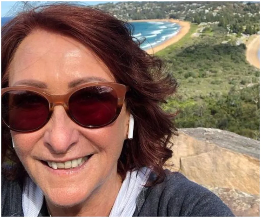 Smiling person with sunglasses and earbuds, overlooking a coastal landscape with a sandy beach and green hills.