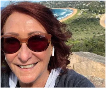 Smiling person with sunglasses and earbuds, overlooking a coastal landscape with a sandy beach and green hills.