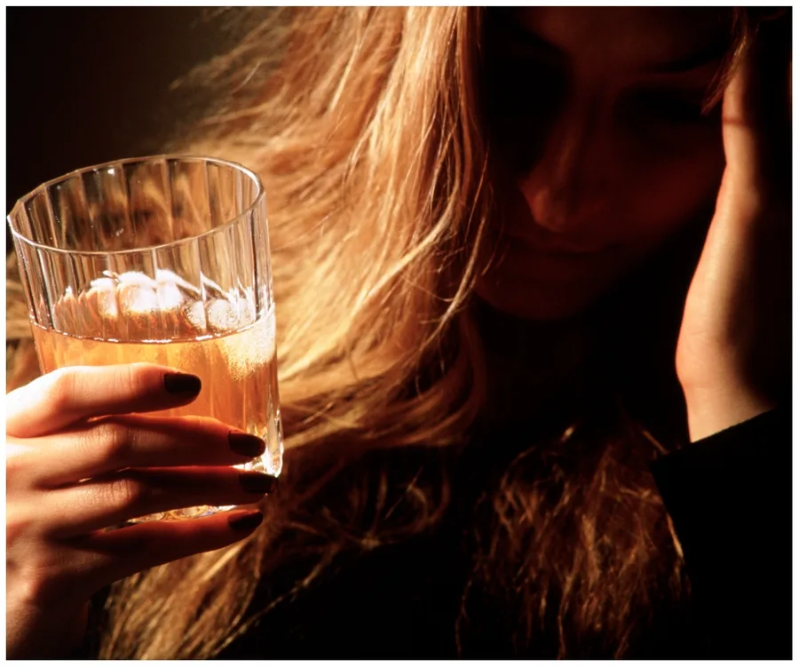 A person with long hair holds a glass of drink, looking contemplative in dim lighting.