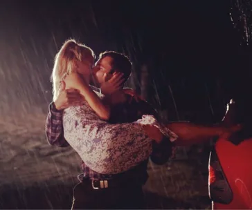 A couple kisses passionately in the rain, the man holding the woman in his arms next to a red car.