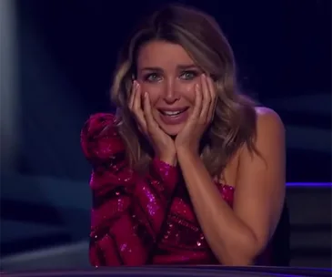 A surprised woman in a glittery red dress holds her face with both hands while sitting on a panel, reacting emotionally.