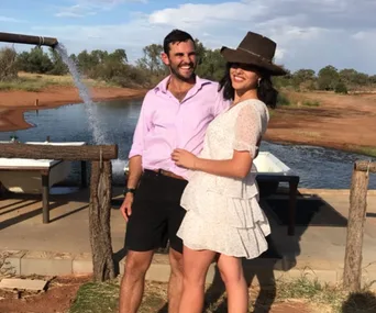 A couple smiling near a water feature; the woman is wearing a white dress and hat, man in a pink shirt and shorts.